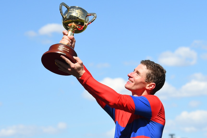 A jockey in Verry Elleegant's blue and red colors smiles as he holds up the Melbourne Cup trophy at Flemington Racecourse.