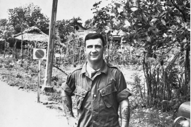 A black and white photo of a smiling man in army fatigues with a Vietnamese village behind him.