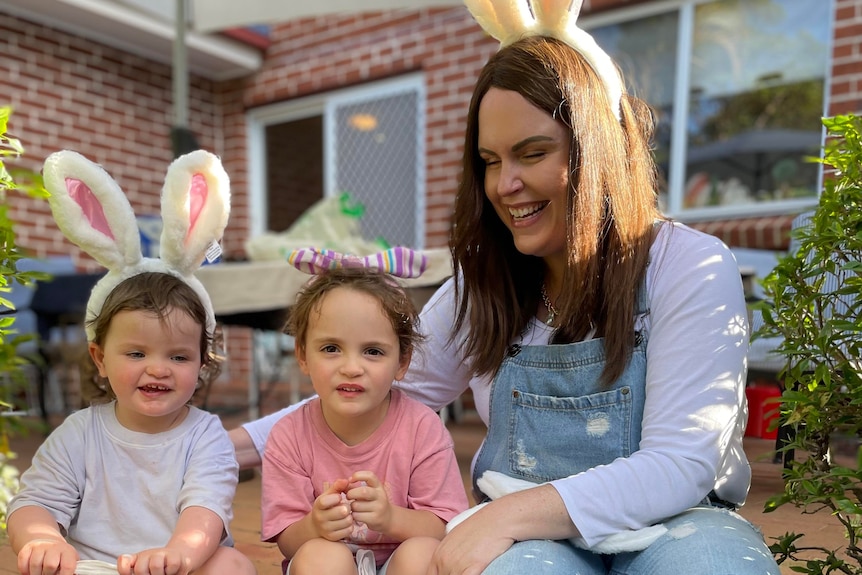 A woman with dark hair sits on a step with two young children, they are all wearing bunny ears