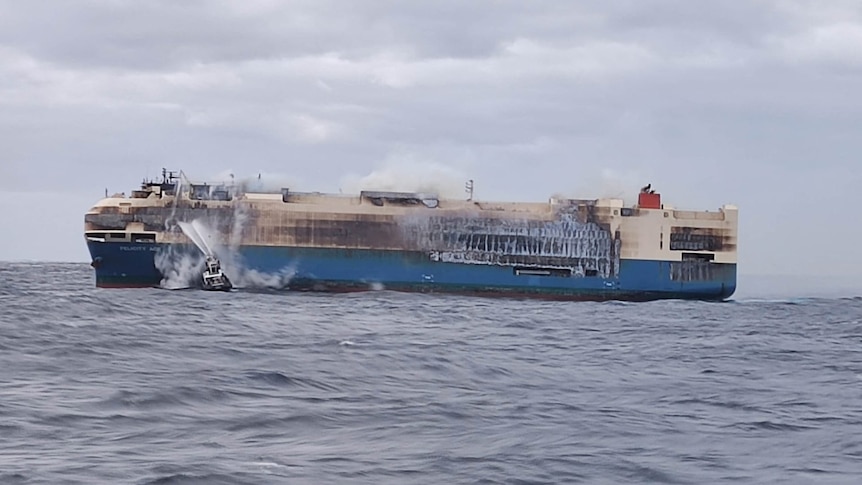 A burnt white cargo ship floats in the middle of the ocean.