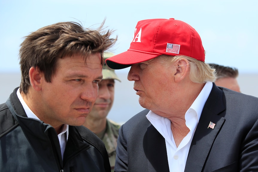 Donald Trump in a red cap whispers in Ron DeSantis's ear 