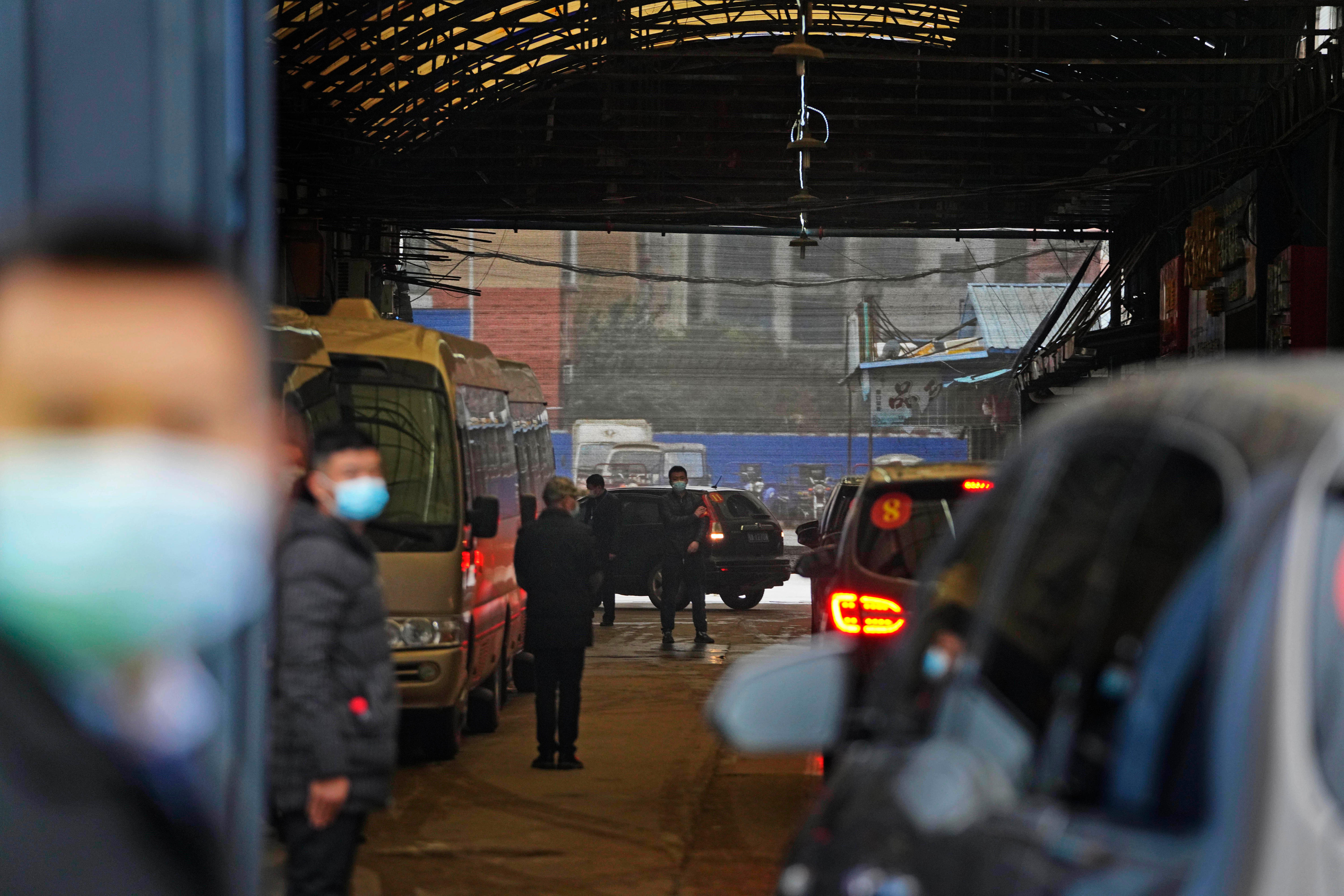 A line of vehicles appears under a large partially exposed structure, masked people stand around