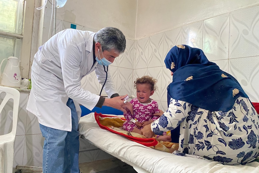 A doctor in a face mask assesses a toddler in a hospital bed