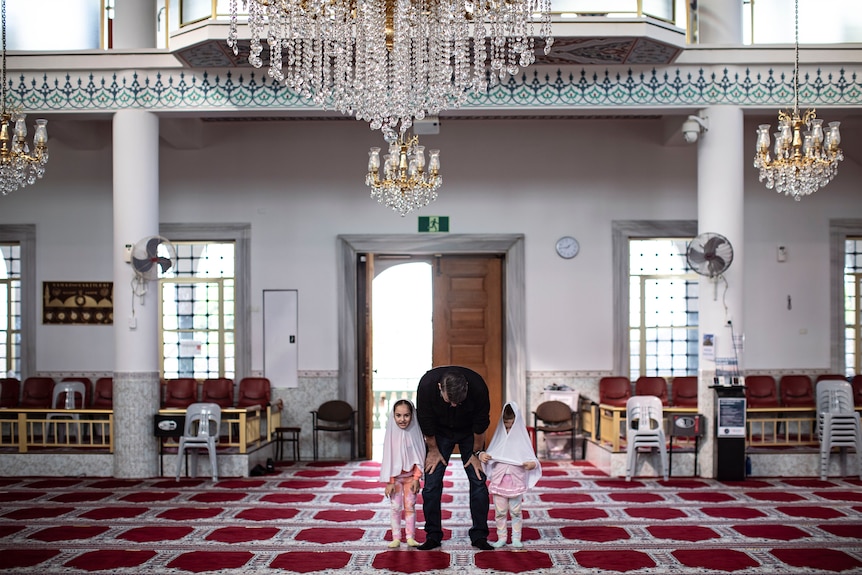 A father bows down inside a mosque, next to his two daughters, both wearing hijabs.