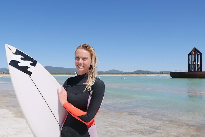 A woman with blonde long hair wears a wetsuit and holds a surfboard on the beach of a wave pool