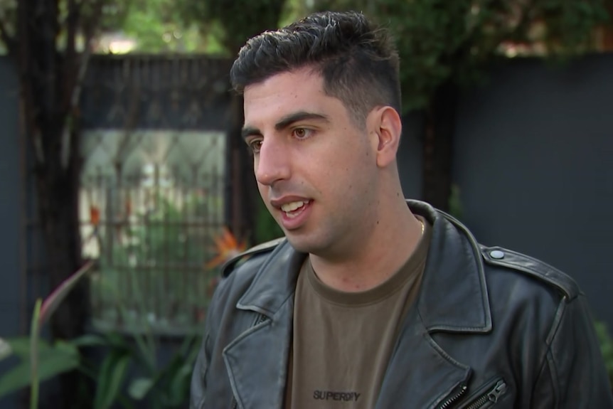 Matthew wears a brown shirt and black leather jacket and stands in the garden of a home.