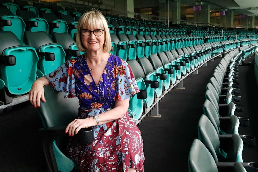Caucasian woman with short blonde hair and glasses wearing a floral dress sitting on a seat within an empty sports stadium.