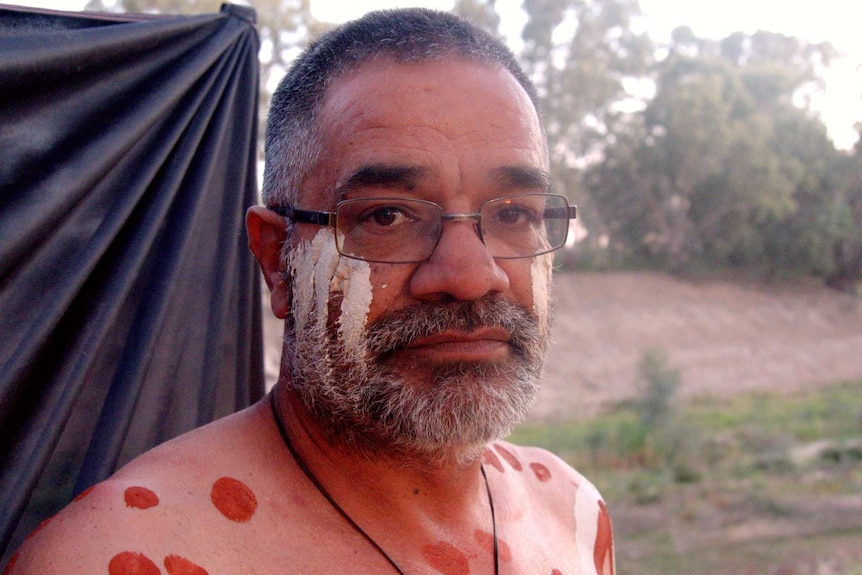 An Aboriginal man in glasses looks down the barrel of the camera.