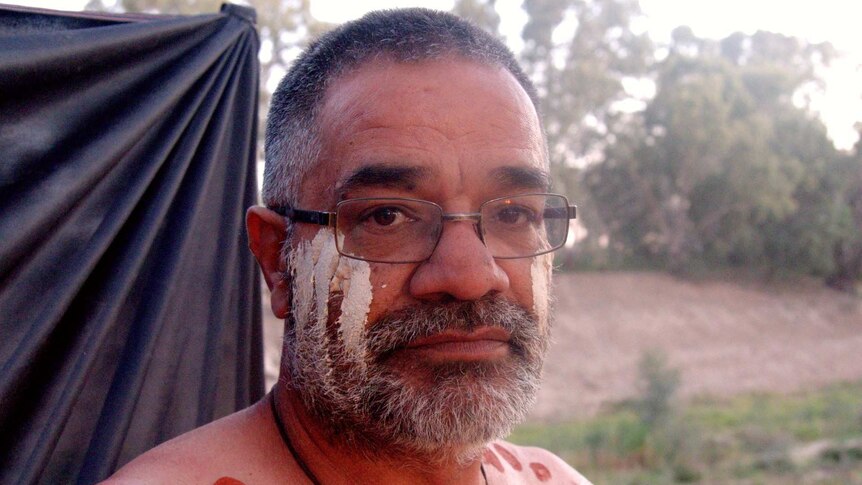 An Aboriginal man in glasses looks down the barrel of the camera.