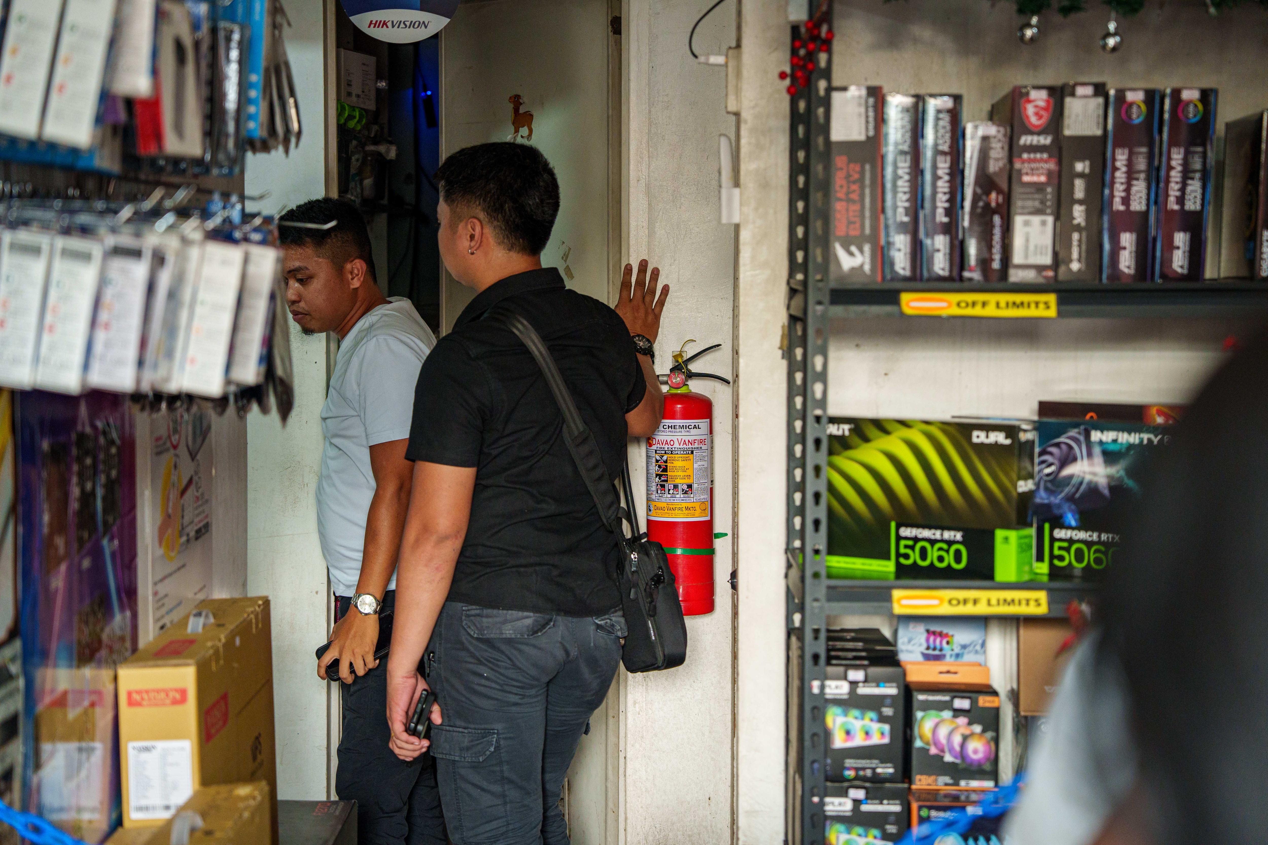Um homem vestindo uma camiseta preta está parado com a palma da mão apoiada na parede. Outro homem vestido está na frente.