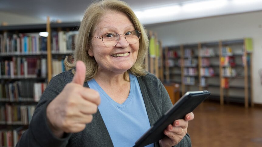 A woman in a blue t shirt is holding a tablet and giving the thumbs up