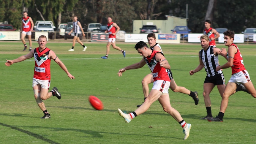 A man in a red, white and black Benalla guernsey kicks a football while people run around him