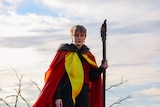 A person wearing a red, black and yellow cloak holds a stick and stands in front of a cloudy blue sky.