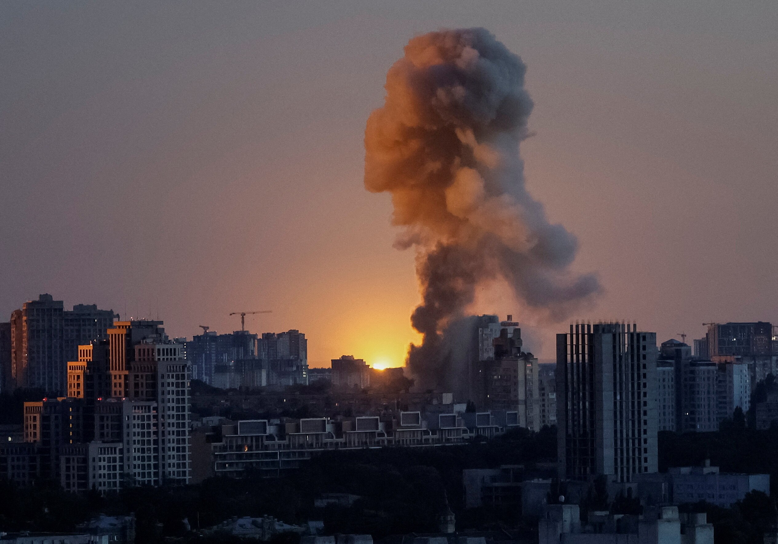 A large plume of smoke rising from the dense skyline of a Kyiv neighbourhood, as the sun rises in the background.