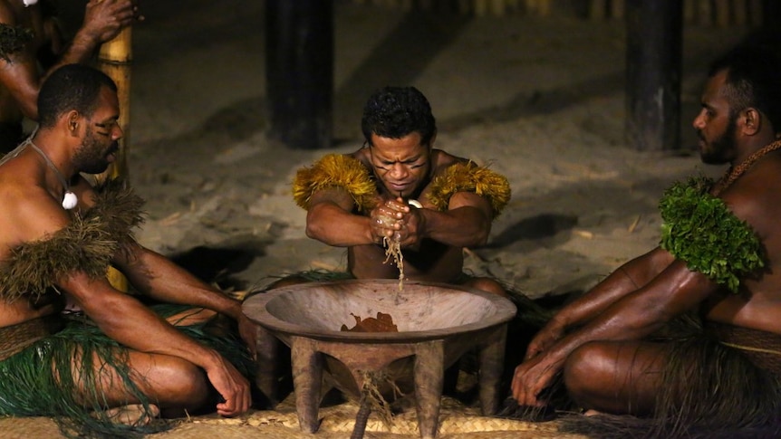Three men sit in grass skirts around a large bowl. The man in the middle squeezes kava juice.