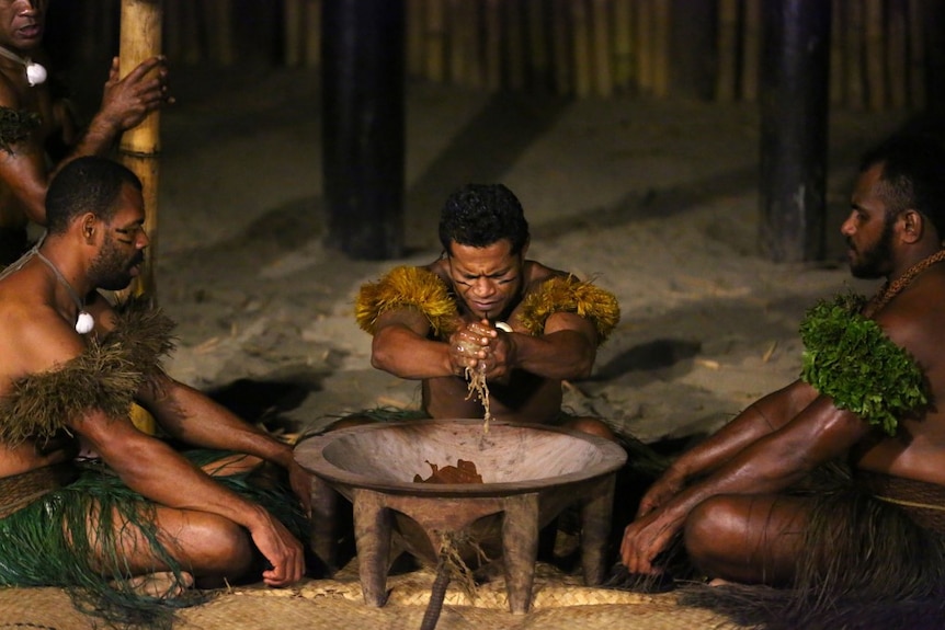 Three men sit in grass skirts around a large bowl. The man in the middle squeezes kava juice.
