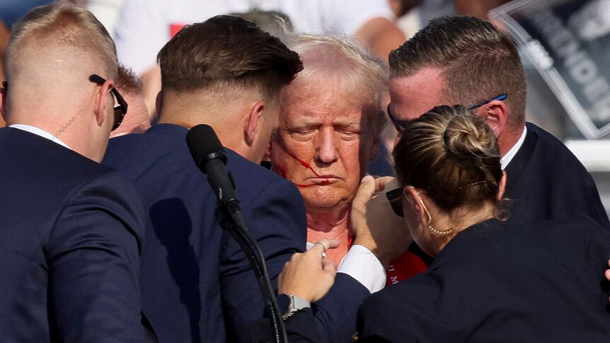 Trump has his eyes closed with a bloody cheek as suited agents surround him