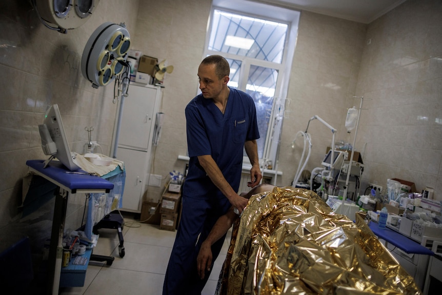 A doctor looks at a screen while treating a man wrapped in gold foil on a hospital bed