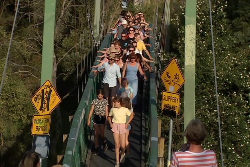 A group of people walk over a suspension bridge near green trees and over a creek.