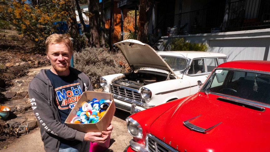 Tim Miller stands in front of his two utes holding a box of plastic lids.