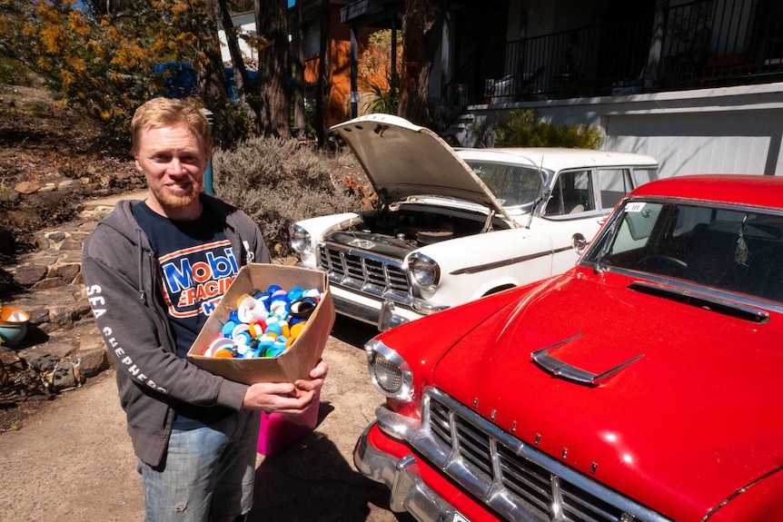 Tim Miller stands in front of his two utes holding a box of plastic lids.