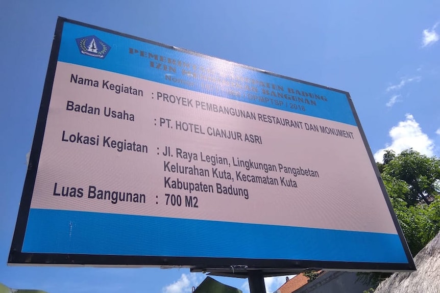 Looking up, a building permit shows plans for a restaurant over one of the two 2002 Bali Bombing sites against a blue sky.