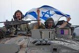 Two armed military personnel looking out of the top of a tank, one holding up an Israeli flag.