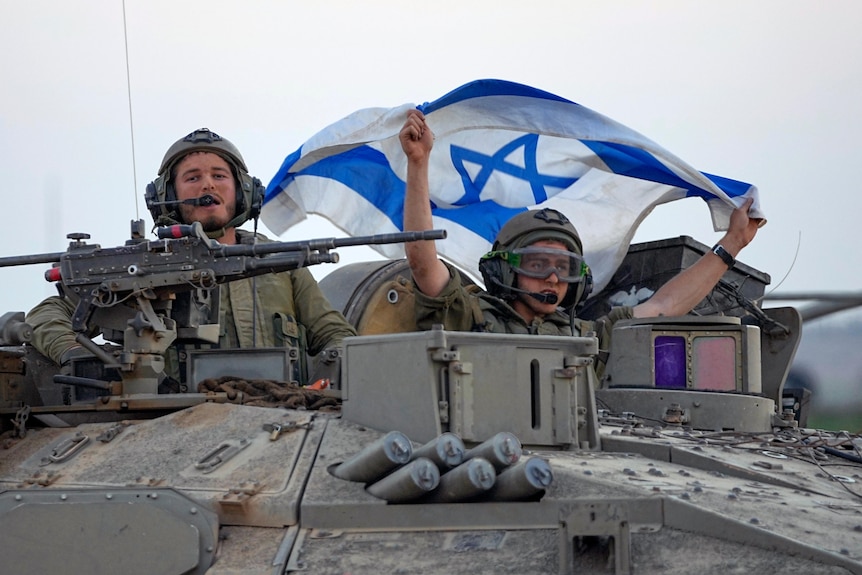 Two armed military personnel looking out of the top of a tank, one holding up an Israeli flag.