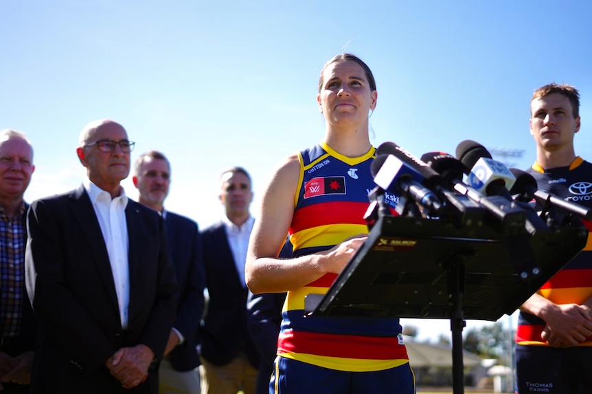 A woman wearing a Crows guernsey stands at a podium with media microphones. A group of men stand behind her