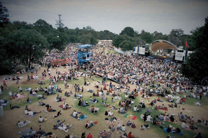 Thousands of people sit around an outdoor stage in a park with trees.