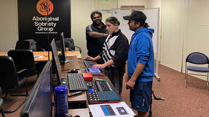 Two men and a teenage boy are standing in front of three computer screens. One man has his hand on the boy's shoulder.