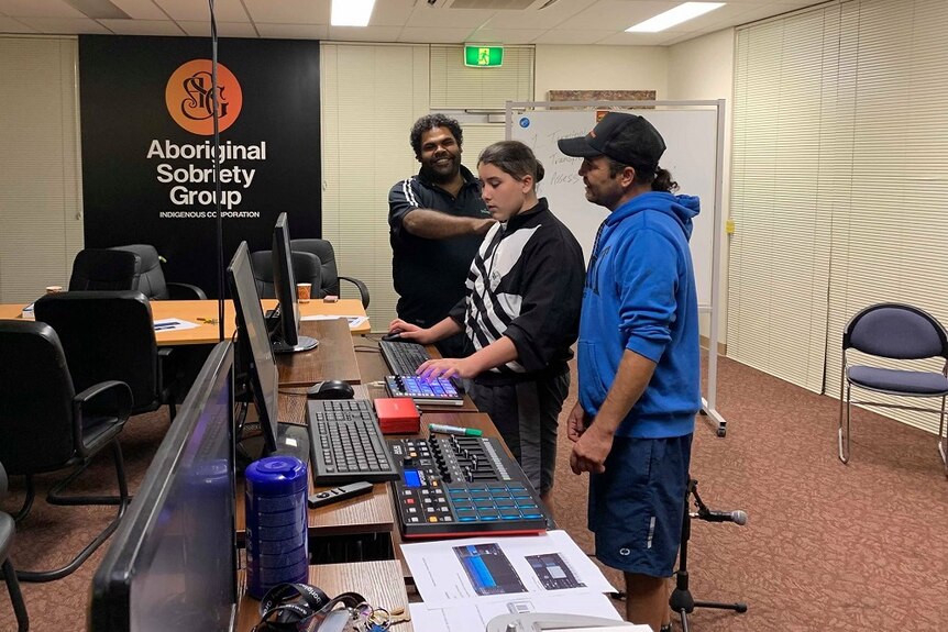 Two men and a teenage boy are standing in front of three computer screens. One man has his hand on the boy's shoulder.