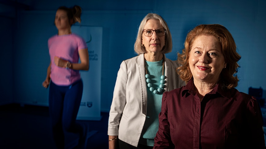 Deirdre McGhee and Julie Steele stand in a lab with a woman running on a treadmill behind them.