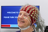 elderly woman sits looking at the camera with a red cap on her head with colour-coded electrodes and wires