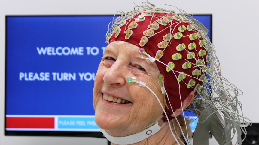 elderly woman sits looking at the camera with a red cap on her head with colour-coded electrodes and wires