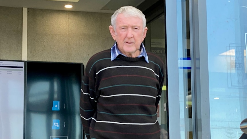 Elderly man standing in front of court sliding doors with hand behind his back wearing striped jumper