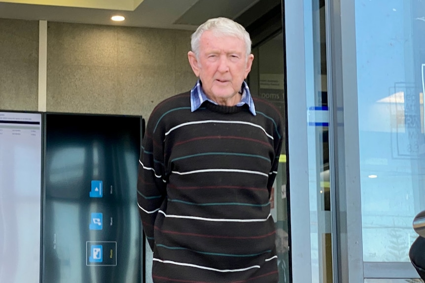 Elderly man standing in front of court sliding doors with hand behind his back wearing striped jumper