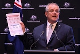 Scott Morrison holds piece of paper in front of a microphone in front of a black background. He is wearing a suit.