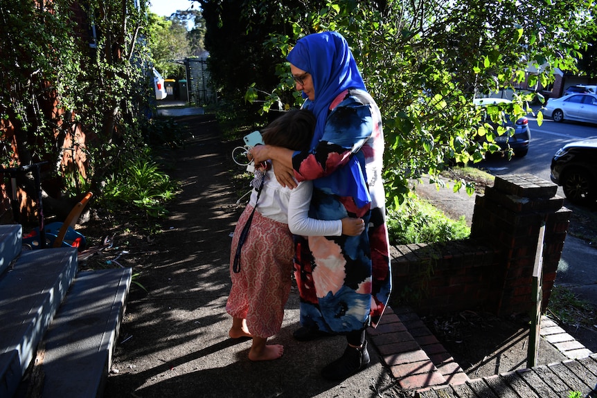 A woman wearing a bright long-sleeve dress and neon blue headscarf as well as glasses hugs a young child in a home frontfootpath