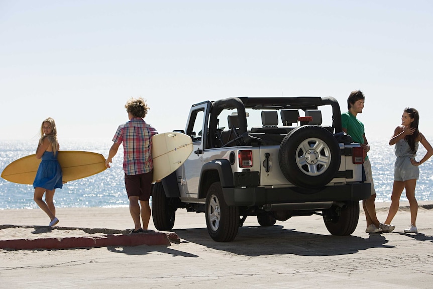 A family of four — two carrying surfboards — stand on a beach next two a four-wheel drive.