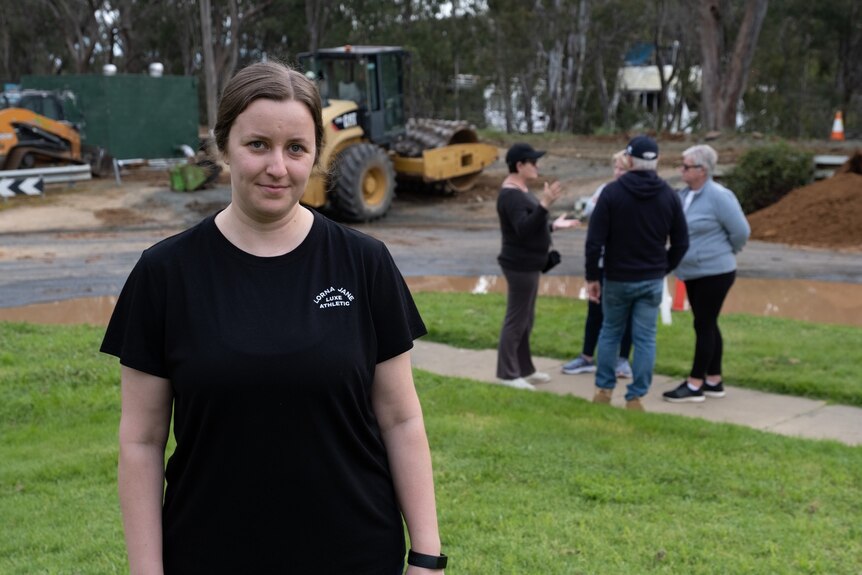 A young woman stands in front of road machinery and flooding.