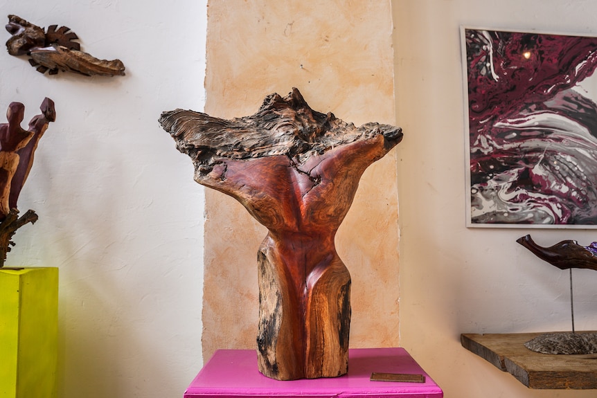 A woman's body shape sculpted into a piece of red wood on a stand against a gallery wall.
