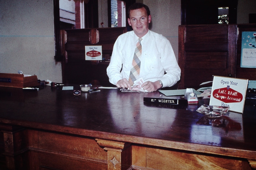 Una foto antigua de un hombre sentado detrás de un escritorio en una oficina bancaria.