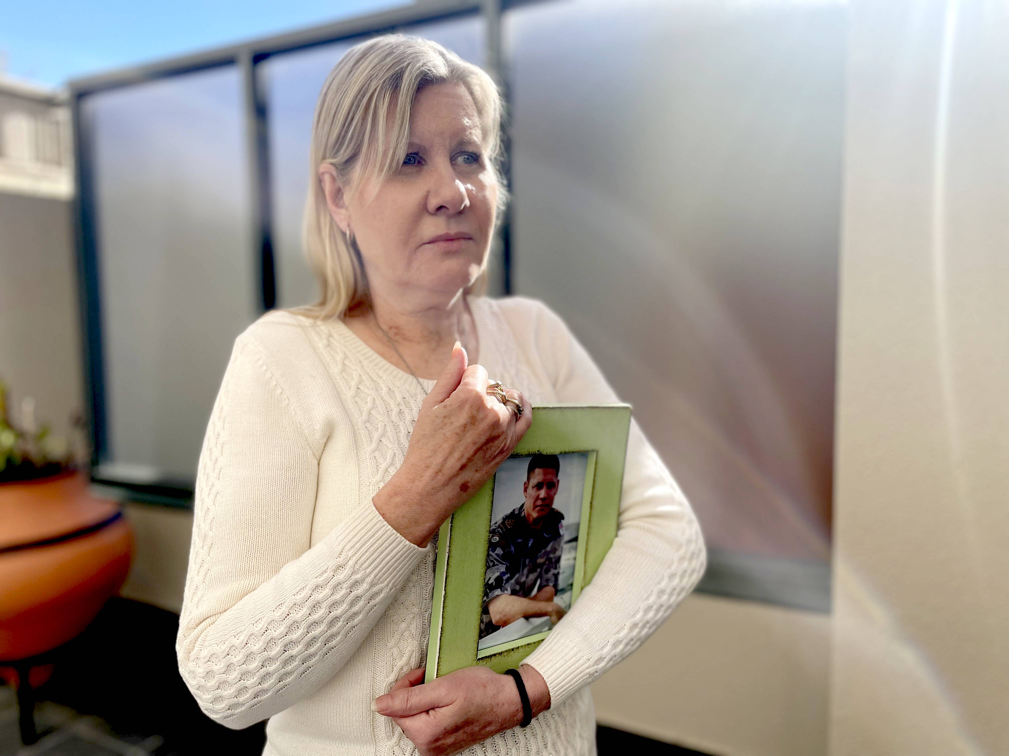 A sad woman in a white knit jumper mother clutches a photography of a young man in army uniform.