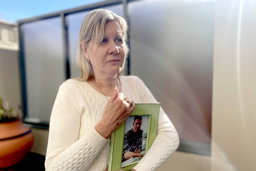 A sad woman in a white knit jumper mother clutches a photography of a young man in army uniform.