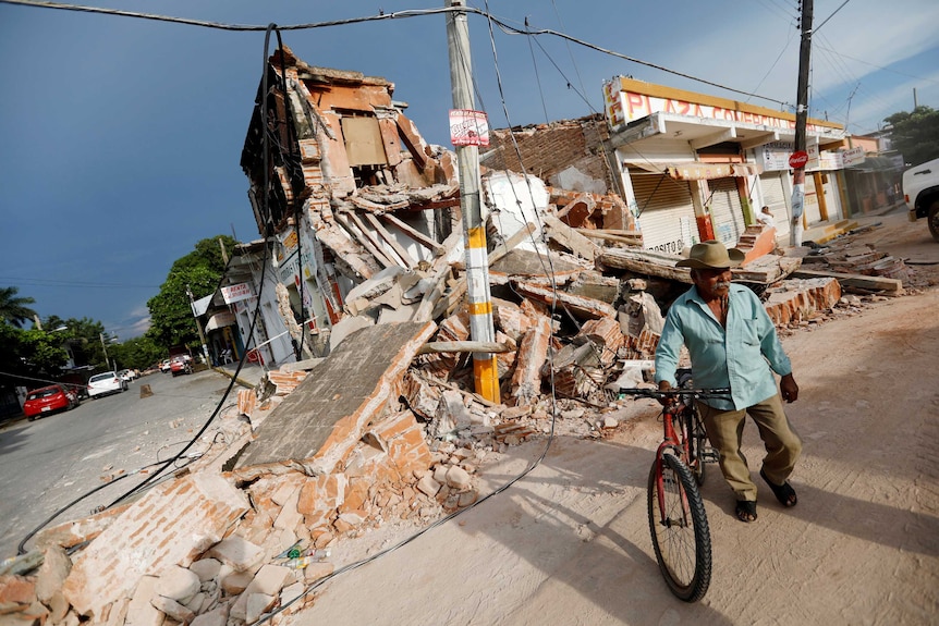 A man with dark skin and a grey moustache stands with a bicycle near the remains of a building.