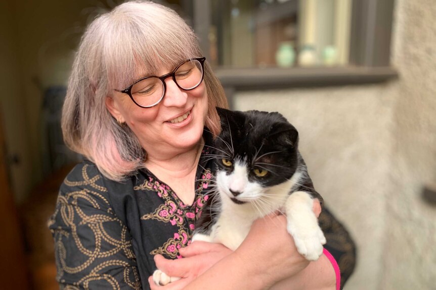 A woman with light-coloured hair and glasses smiles while holding a black and white cat.