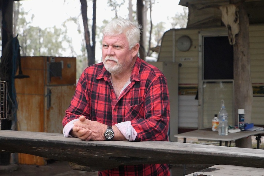 A man in a check red shirt leaning on a timber bench in front of a caravan