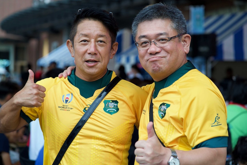 Two men wearing Australian rugby jerseys smile at the camera and give a thumbs up.