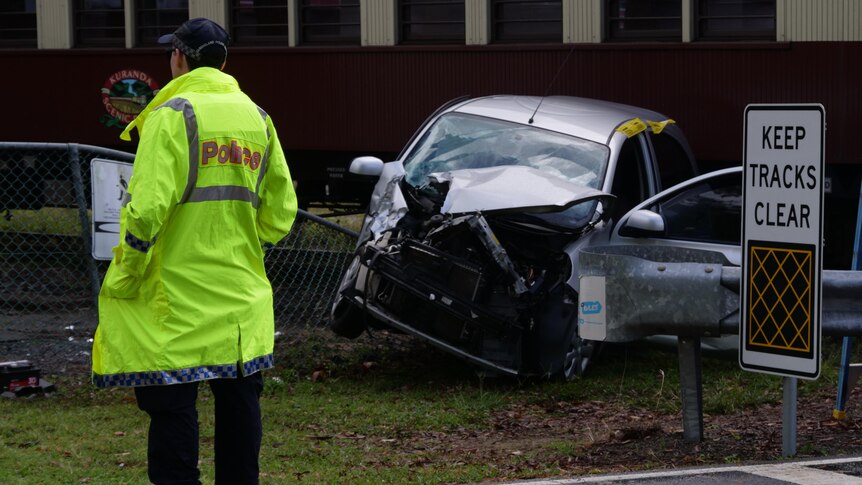 A police officer in a hi-vis jacket stands next to the wreckage of a silver hatchback on a railway track